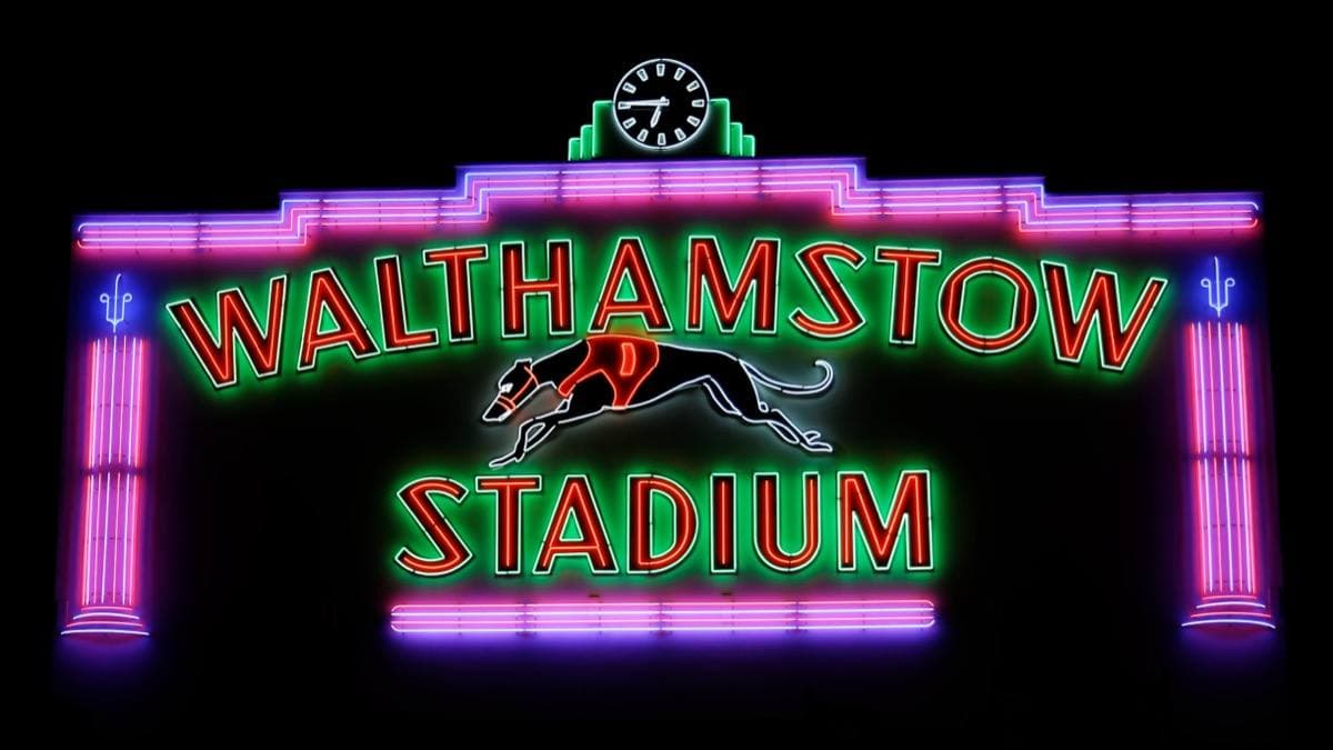 Neon lettering reading Walthamstow Stadium in red and green inside a pink neon frame, a running greyhound between the words, lit against a black night sky