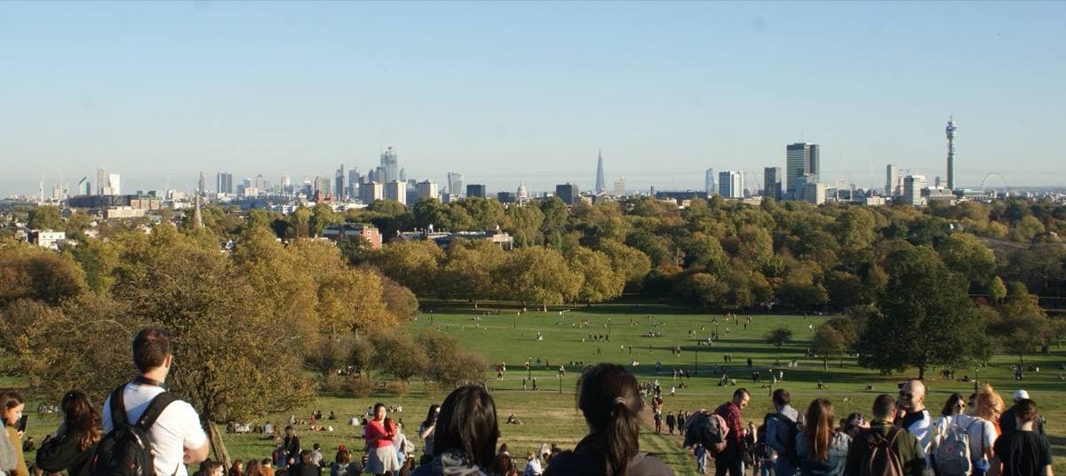 People sitting on the grassy top of Primrose Hill looking out over autumn trees to the London skyline, the BT Tower and the Shard on the horizon