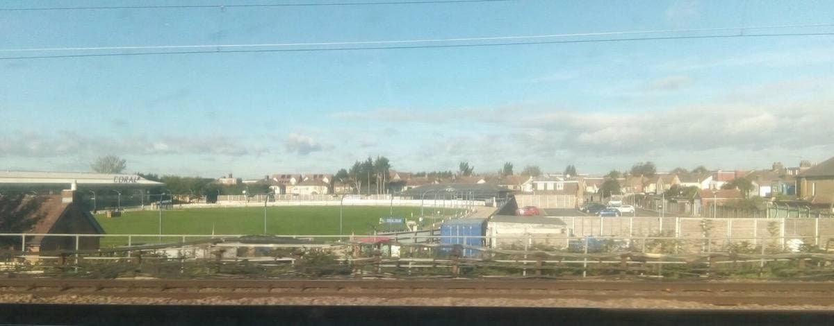 A wide, low view across the green infield and sand track of Romford Greyhound Stadium, stands and houses beyond under a pale sky