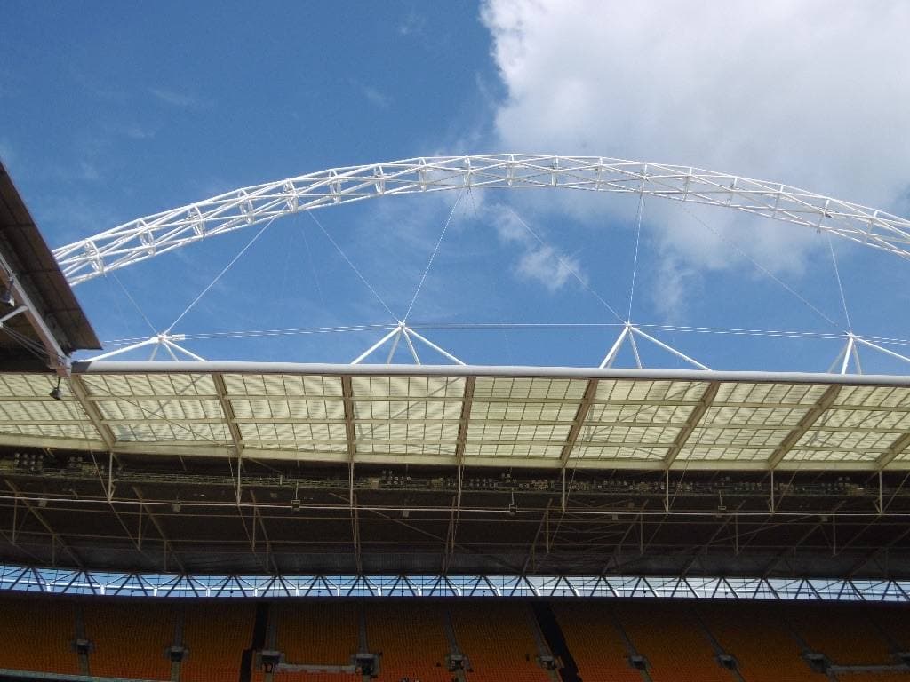 The white steel arch of Wembley Stadium curving high over the stands and roof, seen from inside the stadium against a blue sky