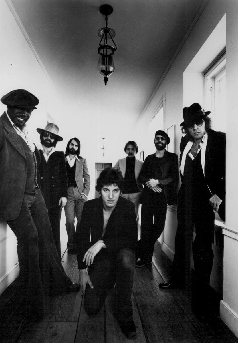 Black and white: the E Street Band standing in a narrow hallway, Springsteen crouching in front, Clarence Clemons on the left