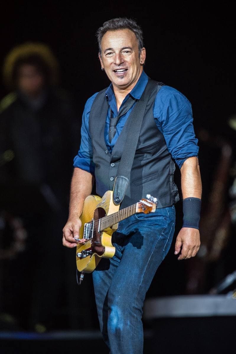 Bruce Springsteen grinning on stage in a blue shirt and dark waistcoat, a worn blonde Fender guitar slung low