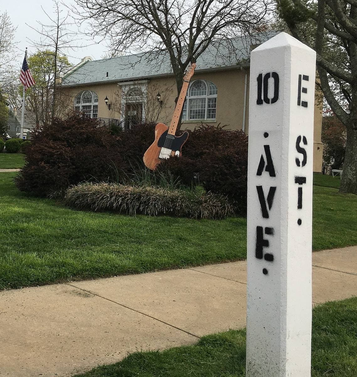 A white wooden post marked 10 Ave and E St on a lawn, with a giant replica of a blonde electric guitar behind it