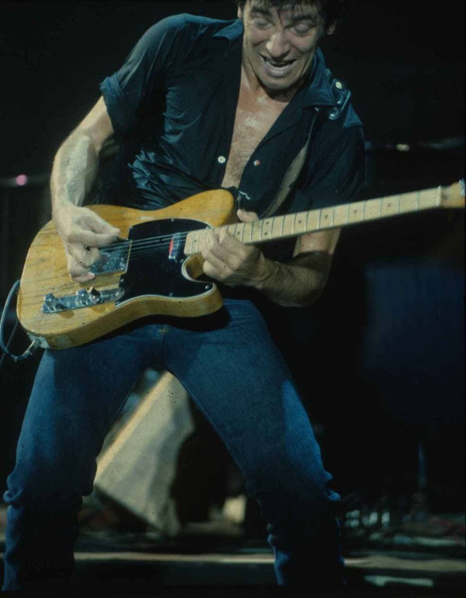 Springsteen in a dark open-necked shirt playing a worn Telecaster, grinning, under stage lights