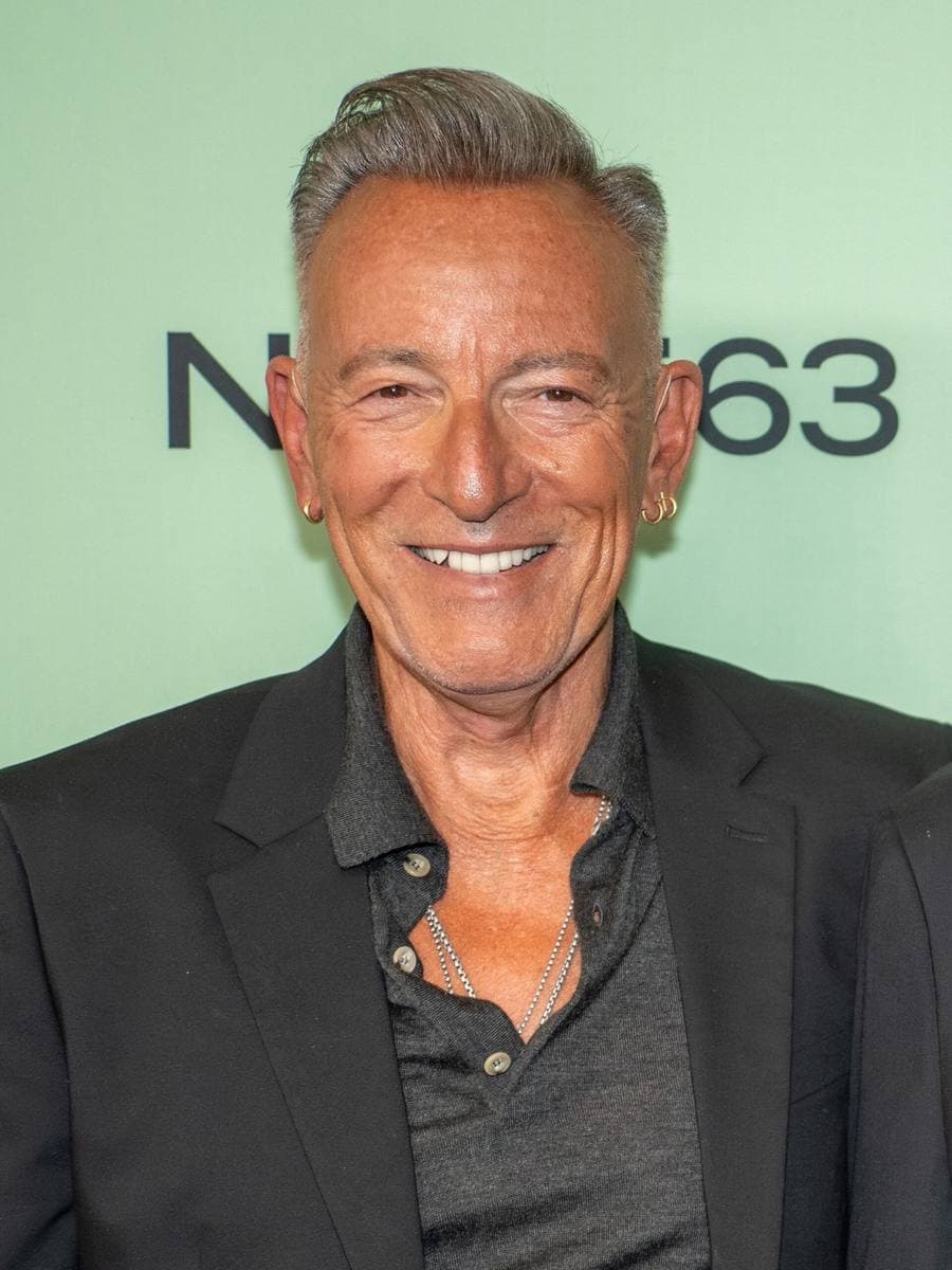 Bruce Springsteen smiling in a dark jacket and open-necked shirt against a pale green backdrop