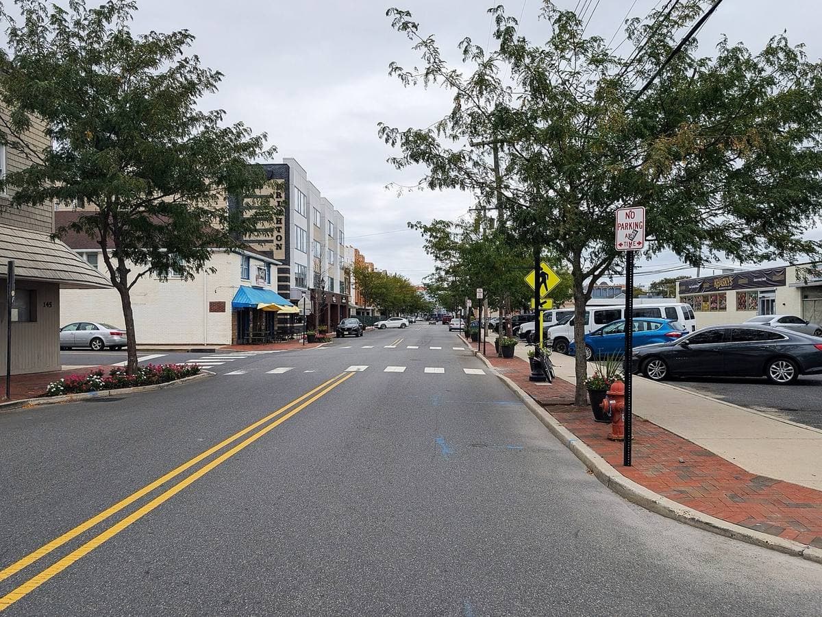 A quiet two-lane town street with shops, trees and parked cars under a grey sky
