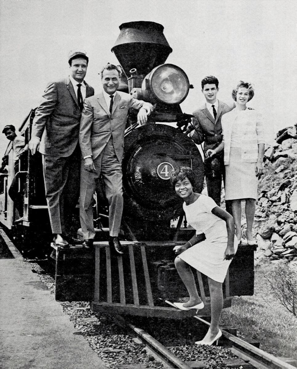 Three men in suits and two young women posed on the front of a small steam locomotive, Little Eva crouching on the buffer