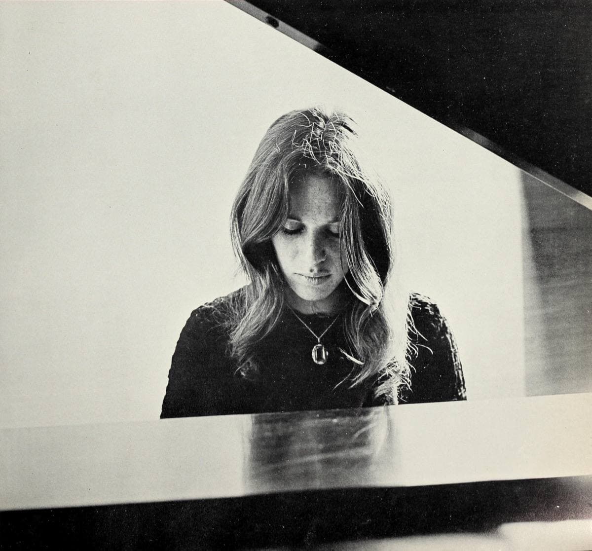 Carole King, long-haired, looking down at the keys of a grand piano with its lid raised