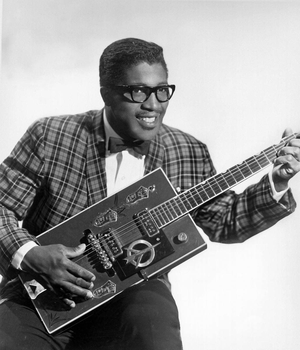 Bo Diddley seated with a rectangular electric guitar