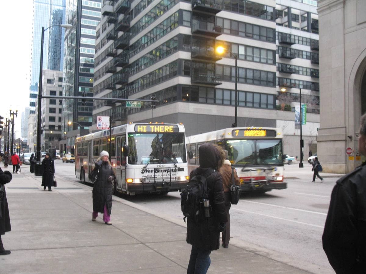 Commuters at a bus stop on a West Loop street corner, two city buses and tall buildings behind