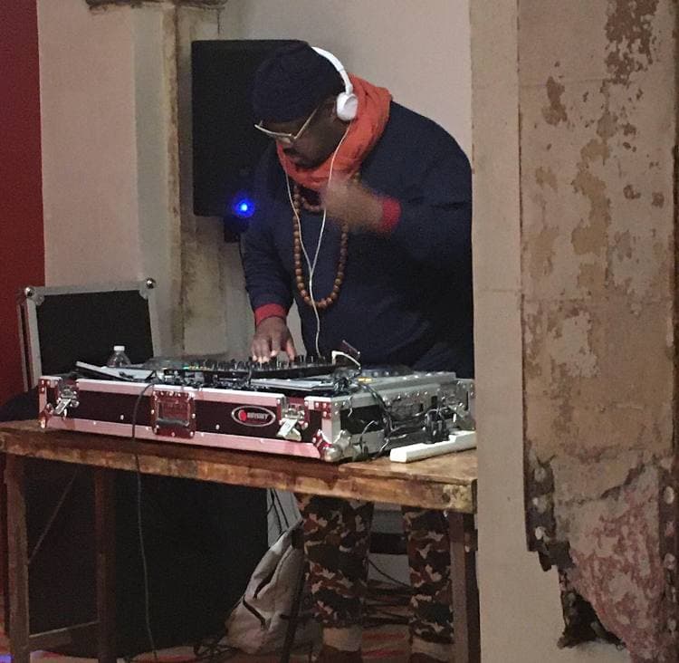 A DJ in headphones and an orange scarf at two turntables on a wooden table