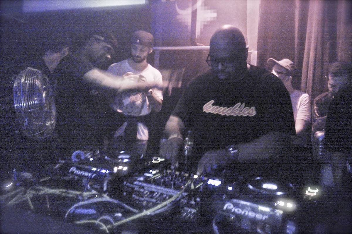Frankie Knuckles in a black T-shirt at a DJ mixer under purple club light, friends behind him