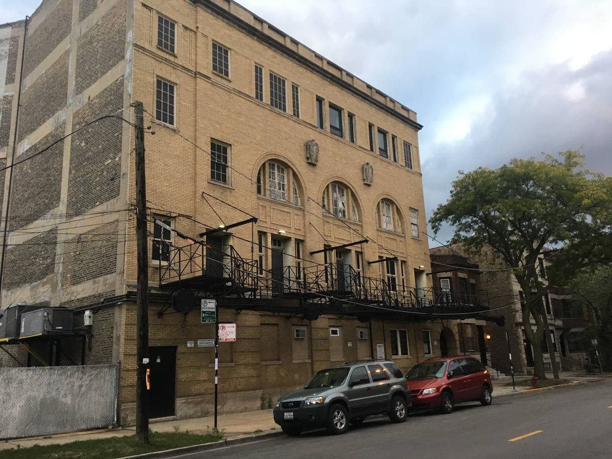 A pale brick former lodge building with arched windows, carved emblems and an iron fire escape, cars parked below