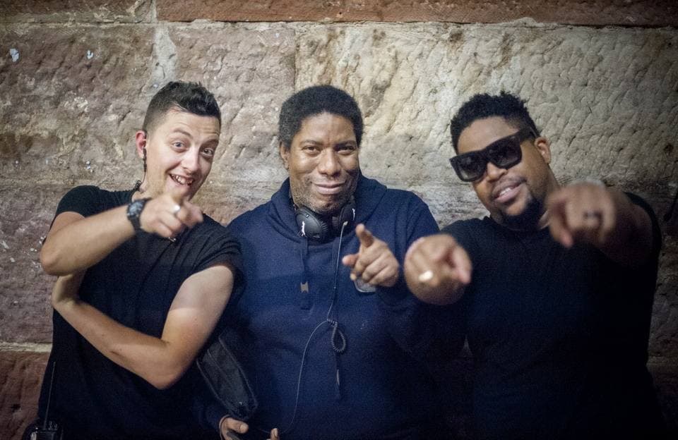 Three men against a sandstone wall pointing at the camera; Marshall Jefferson in the middle in a navy hoodie with headphones round his neck