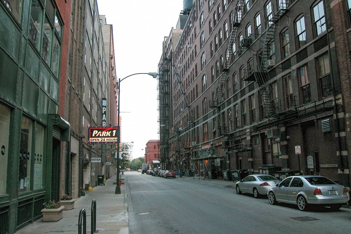 A narrow Printer’s Row street of tall brick loft buildings with fire escapes, a parking sign and parked cars