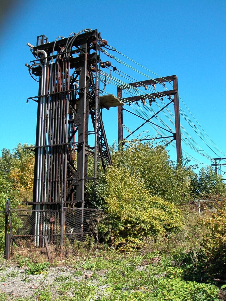 A rusting steel rail tower among weeds under a clear blue sky