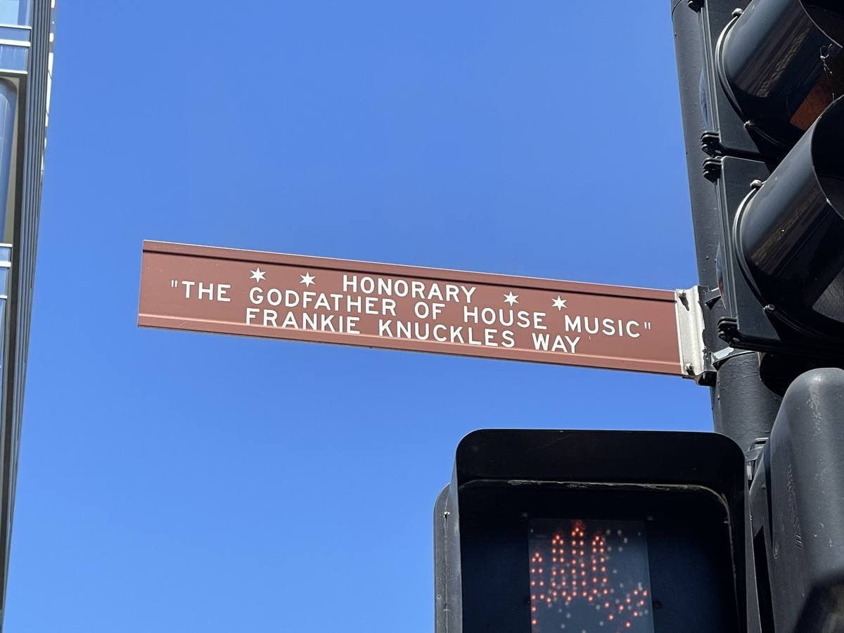 A brown honorary street sign reading The Godfather of House Music Frankie Knuckles Way against blue sky, a pedestrian signal below
