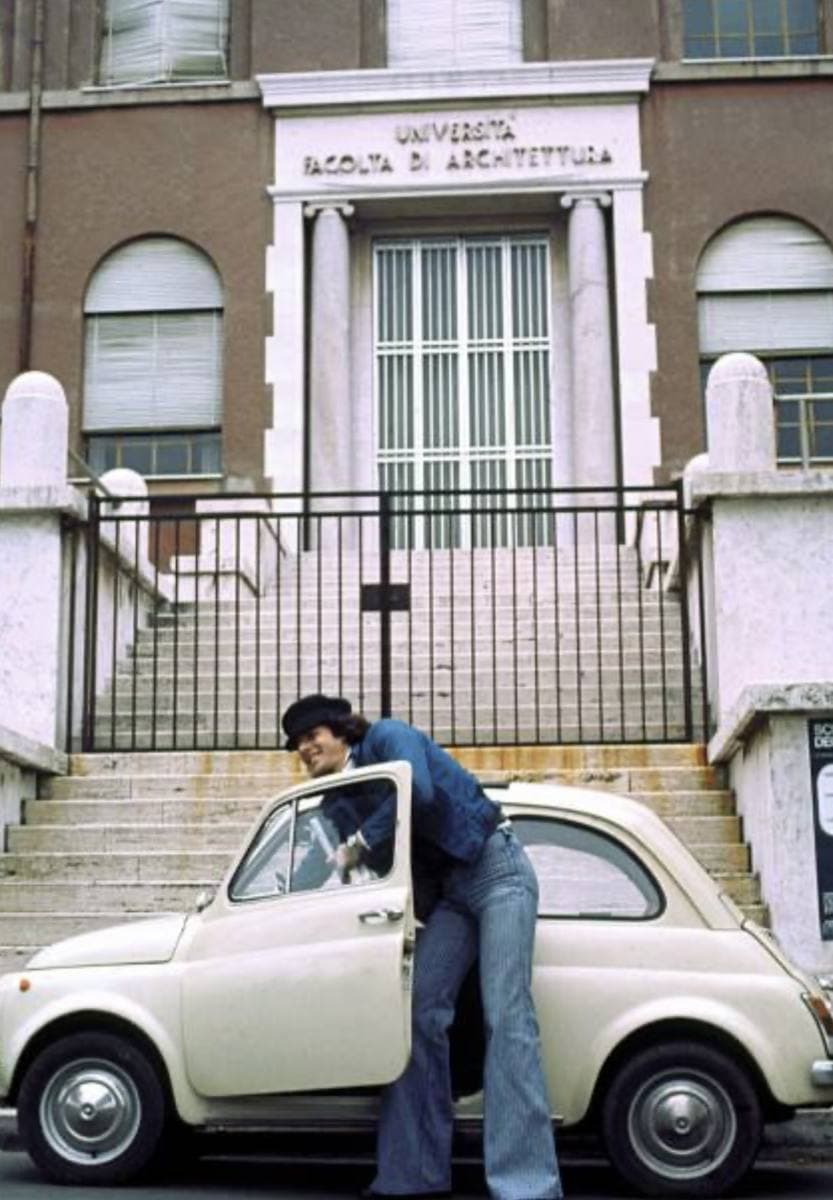 Claudio Baglioni in jeans opening the door of a cream Fiat 500 parked at the foot of the steps of a brick university building