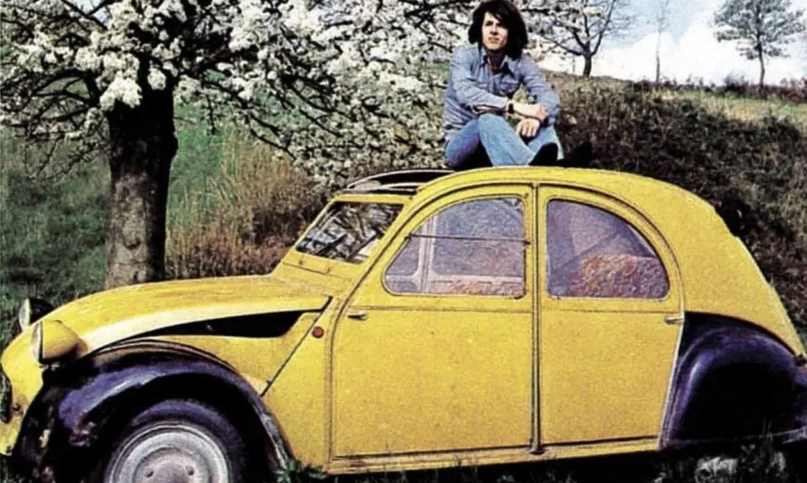 Claudio Baglioni sitting on the roof of a yellow and black Citroën 2CV parked on a grassy slope under a blossoming tree