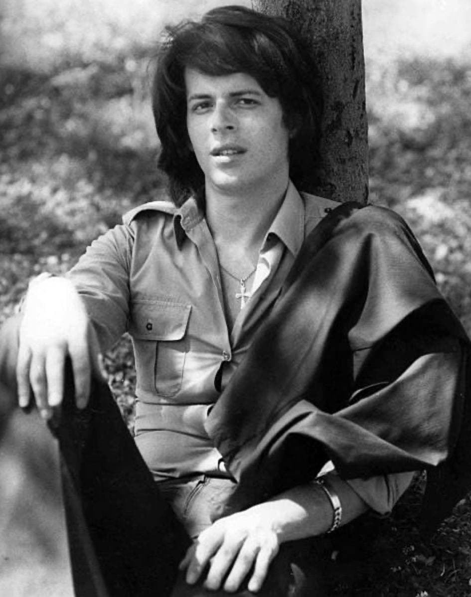 Claudio Baglioni sitting against a tree trunk in a dark shirt, black and white