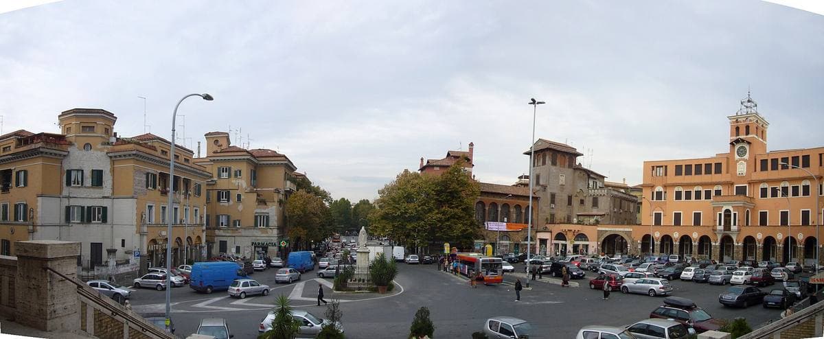 A wide square of ochre and red buildings with a clock tower, cars parked in the centre