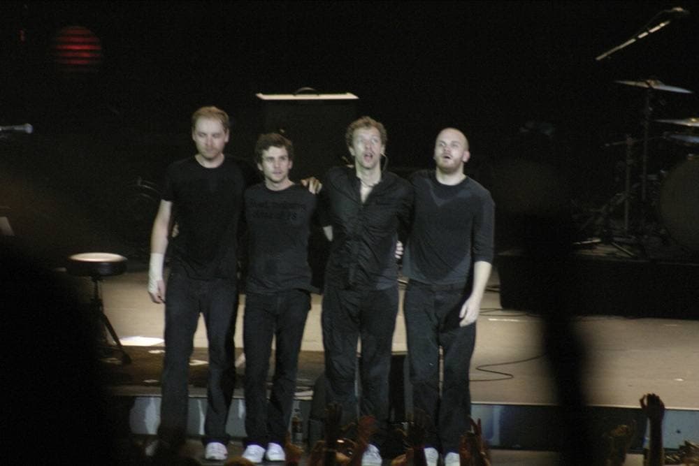 Four men in black standing arm in arm on a dark stage
