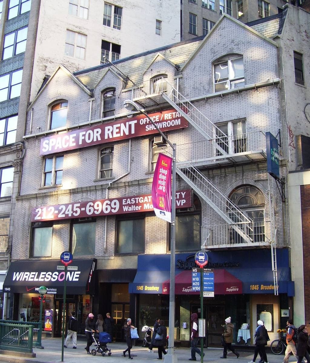A narrow old gabled building with fire escapes and shopfronts on Broadway