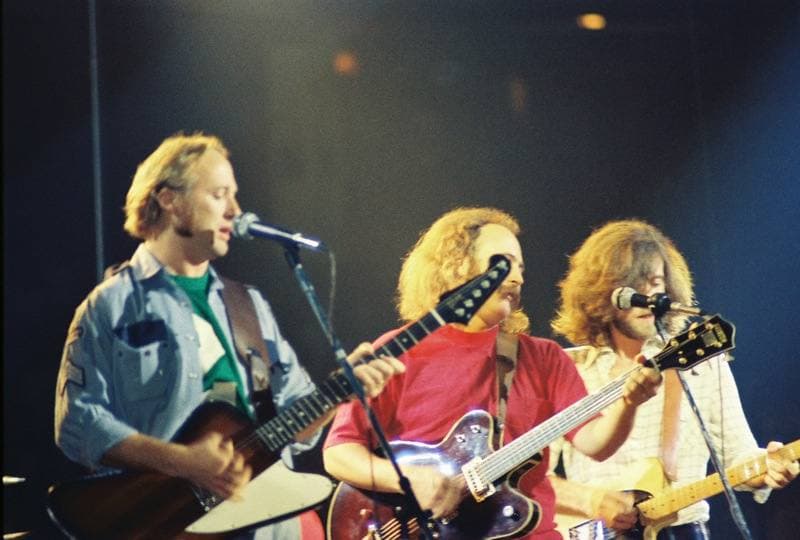 Three musicians in a row at microphones on a dark blue stage, playing electric guitars