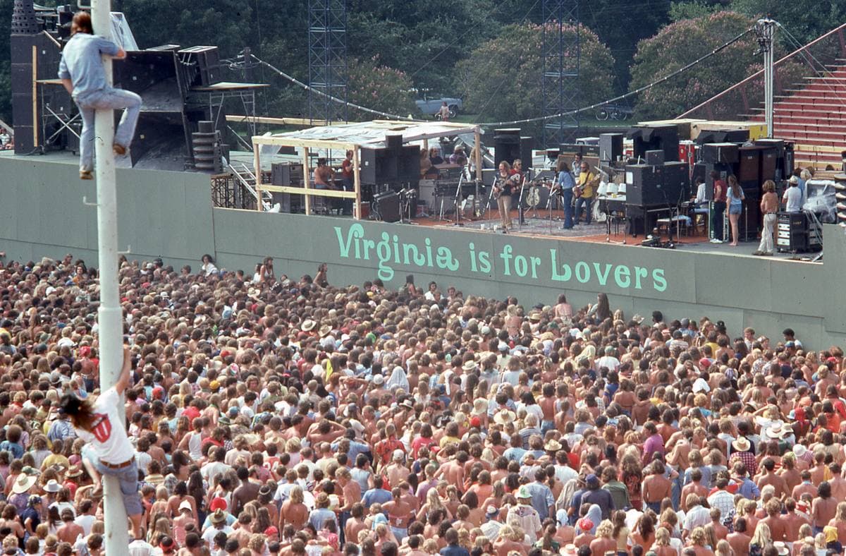 A packed stadium crowd in summer sun facing a stage heaped with speakers, a fan clinging to a pole in the foreground