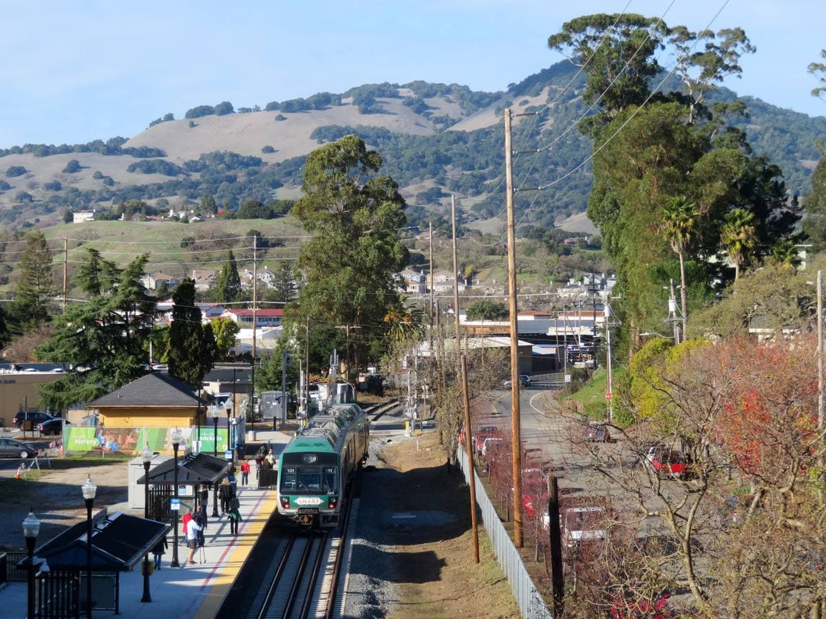 A green hillside rising behind a small railway station and a train in Novato