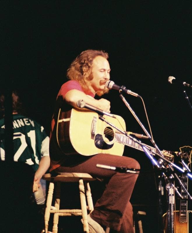 A man with long curly hair and a moustache sits on a wooden stool singing into a microphone with an acoustic guitar