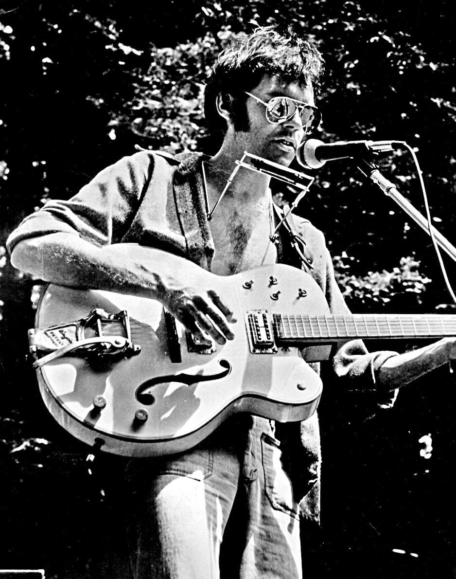 Black-and-white photograph of Neil Young in aviator glasses singing at a microphone with a pale hollow-body guitar