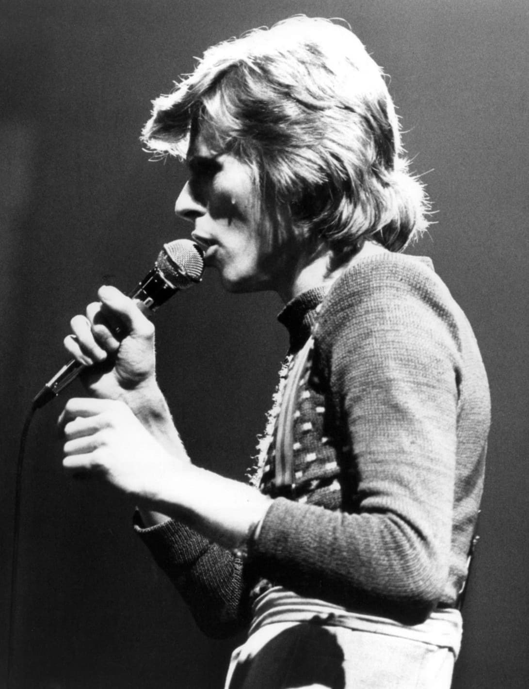 David Bowie sings into a microphone under stage light