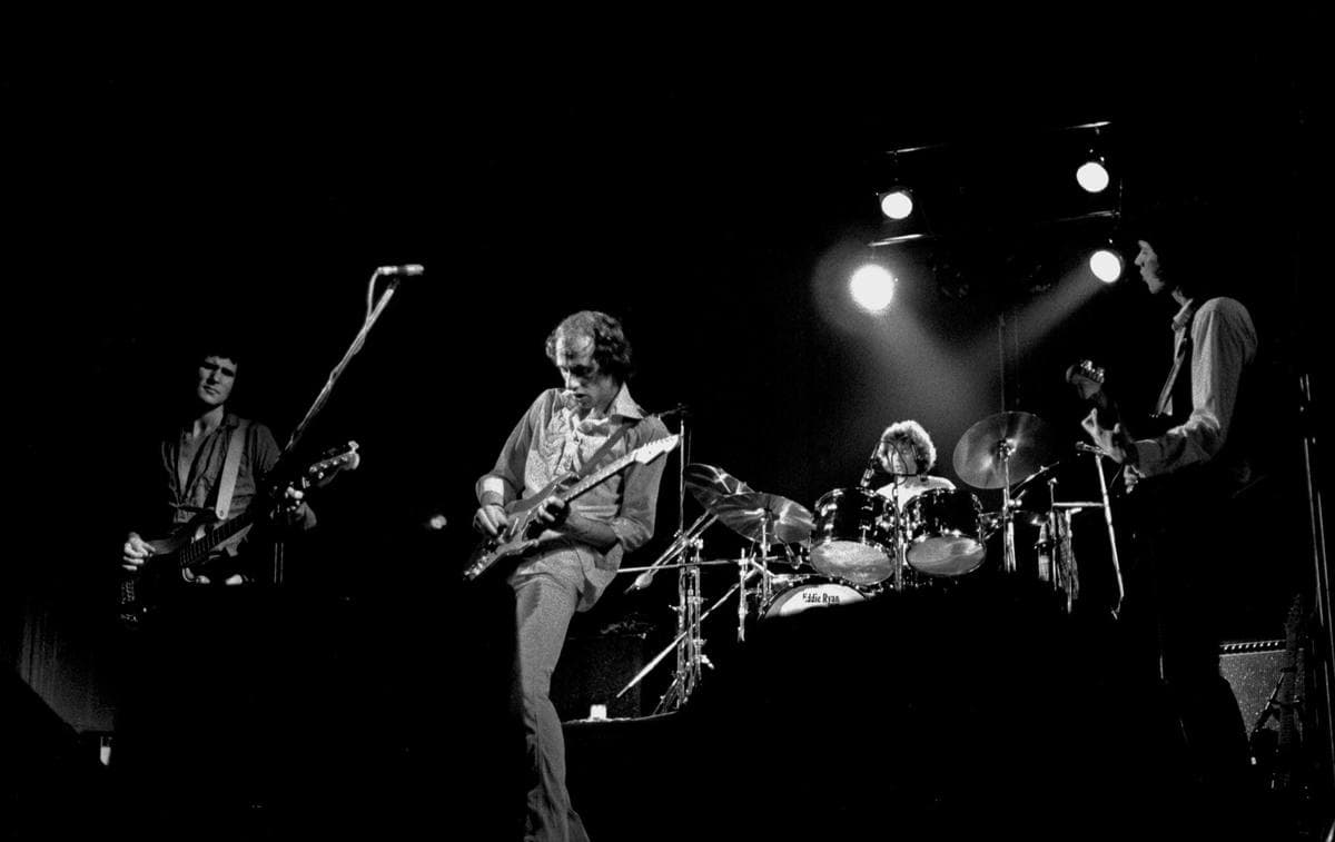 The four original members of Dire Straits on a dark stage under spotlights, black and white