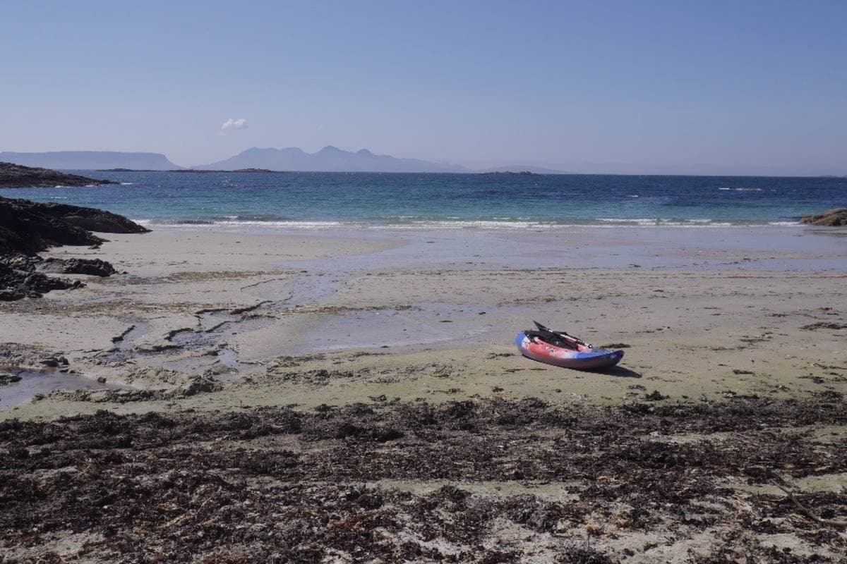 A pale sandy beach and rocks with a kayak, the sea and island hills on the horizon