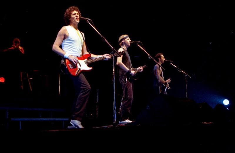John Illsley with a red bass, Mark Knopfler in a headband and a third guitarist at microphones on a dark stage