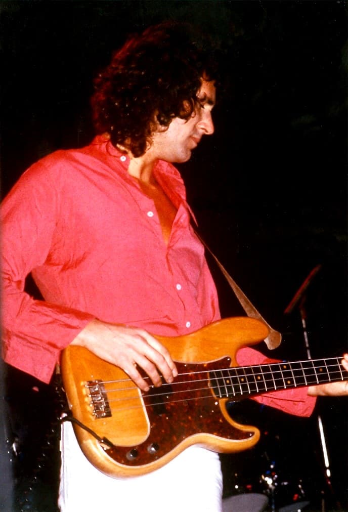 John Illsley in a red shirt playing a natural-wood bass guitar on stage