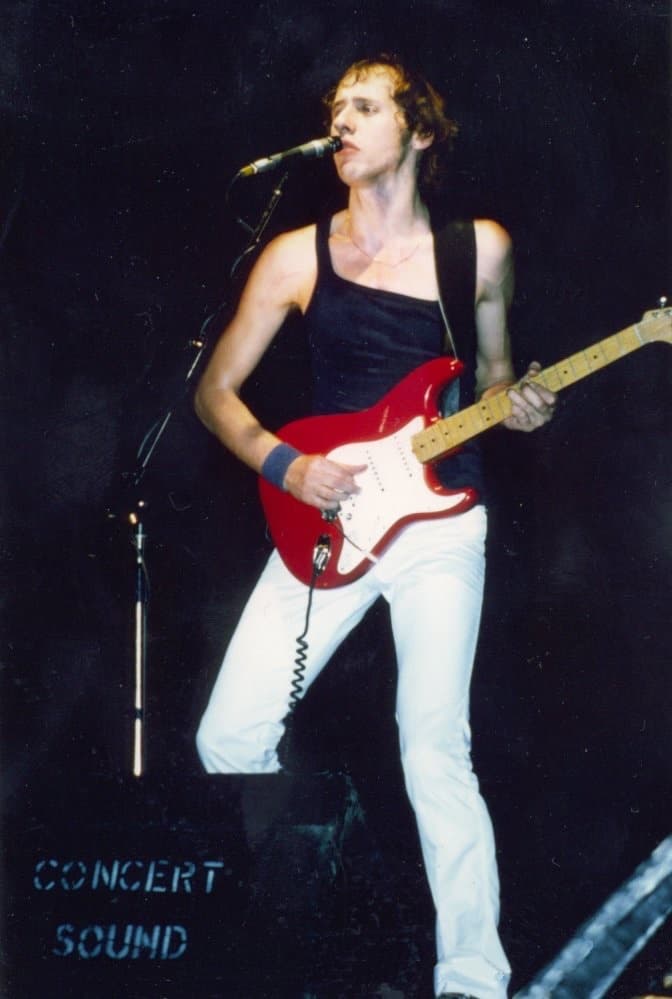 Mark Knopfler in a black vest and white trousers, singing and playing a red electric guitar