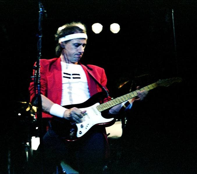 Mark Knopfler in a red jacket and white headband playing a black and white guitar