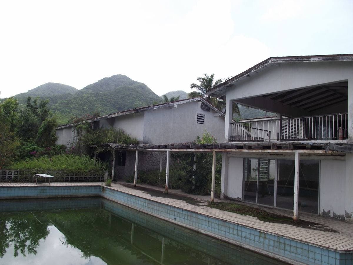 An abandoned white studio building with a green, stagnant swimming pool in front and hills behind