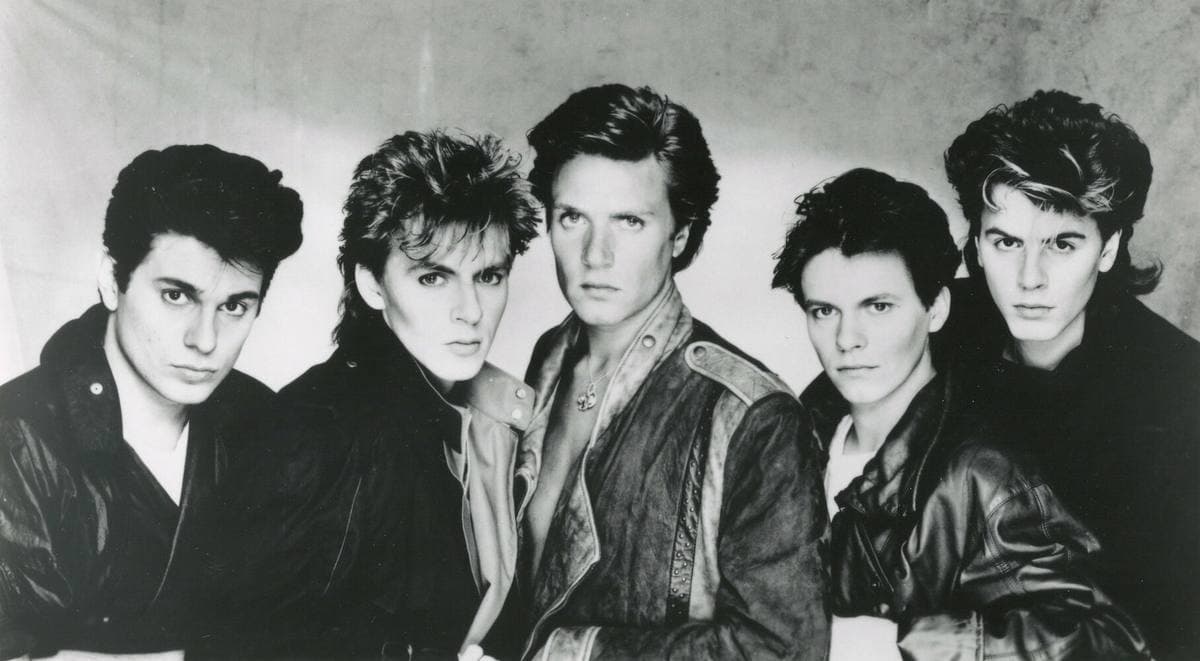 The five members of Duran Duran in 1983, standing in a row in pale suits and loose shirts, hair swept up