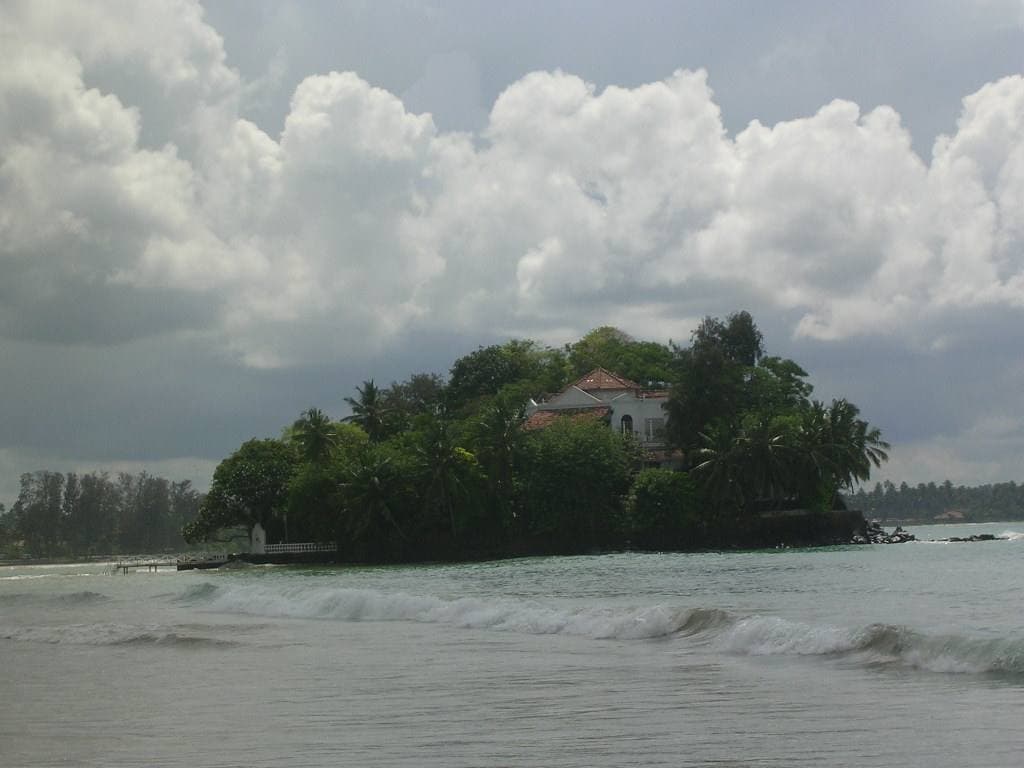 A small wooded island with a white house, just off a sandy beach