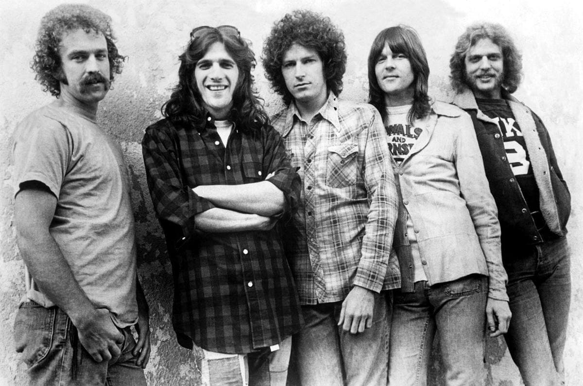Five long-haired young men in denim and check shirts leaning against a rough plaster wall, Glenn Frey with sunglasses pushed up and arms folded