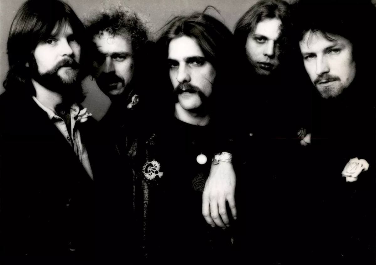 Five bearded and moustached men in dark clothes crowded together against a grey backdrop, a rose pinned to one jacket