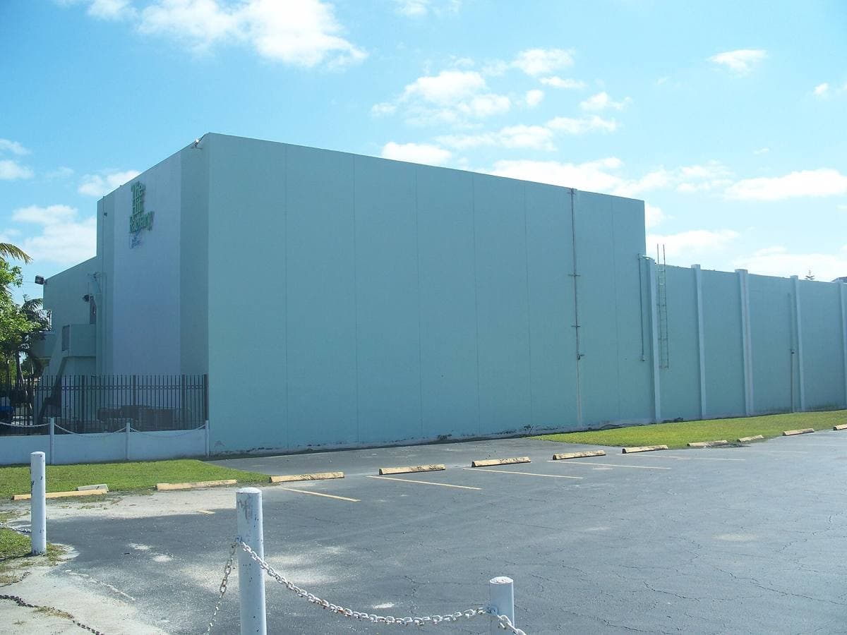 A tall pale-green windowless studio block beside a car park under a blue sky