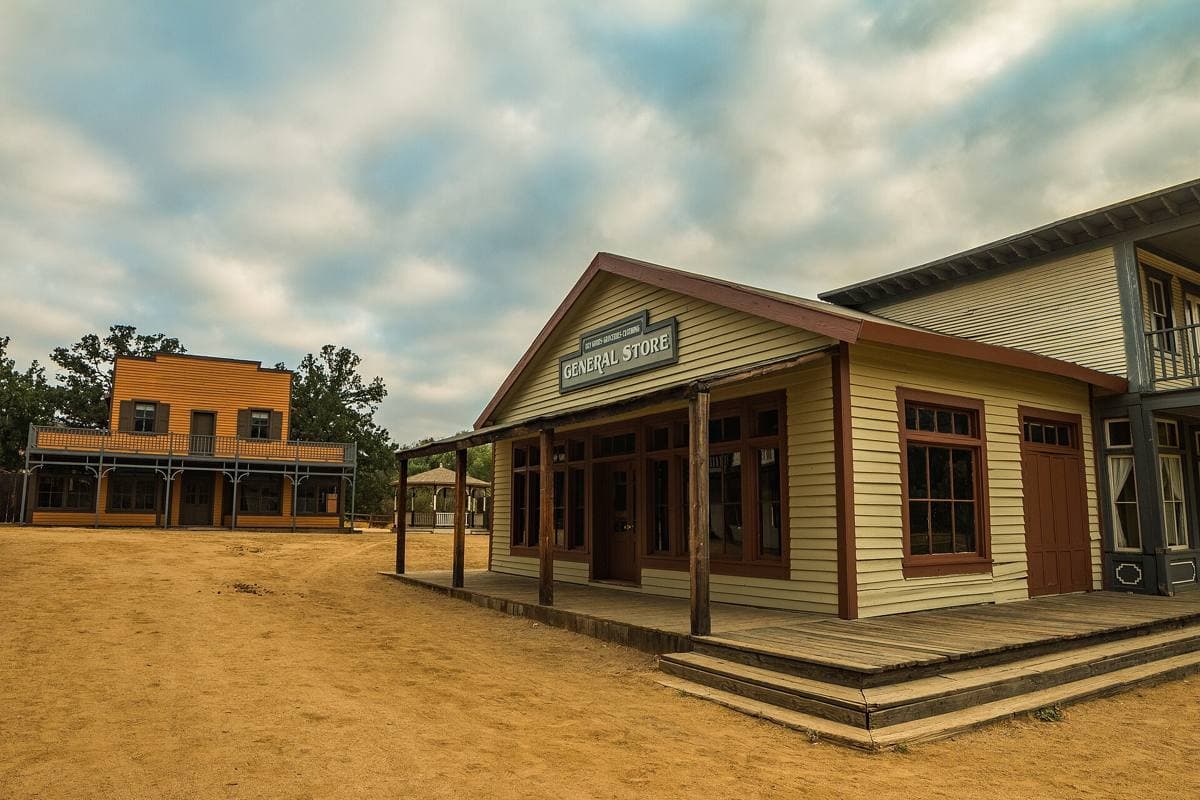 Wooden false-front buildings of a Western film town on a dirt street under oak-covered hills