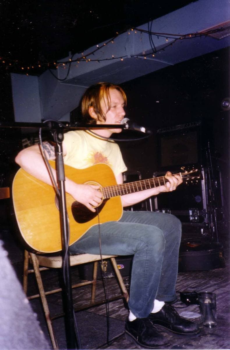 Elliott Smith seated on a folding chair, singing into a microphone and playing an acoustic guitar, fairy lights along a low ceiling