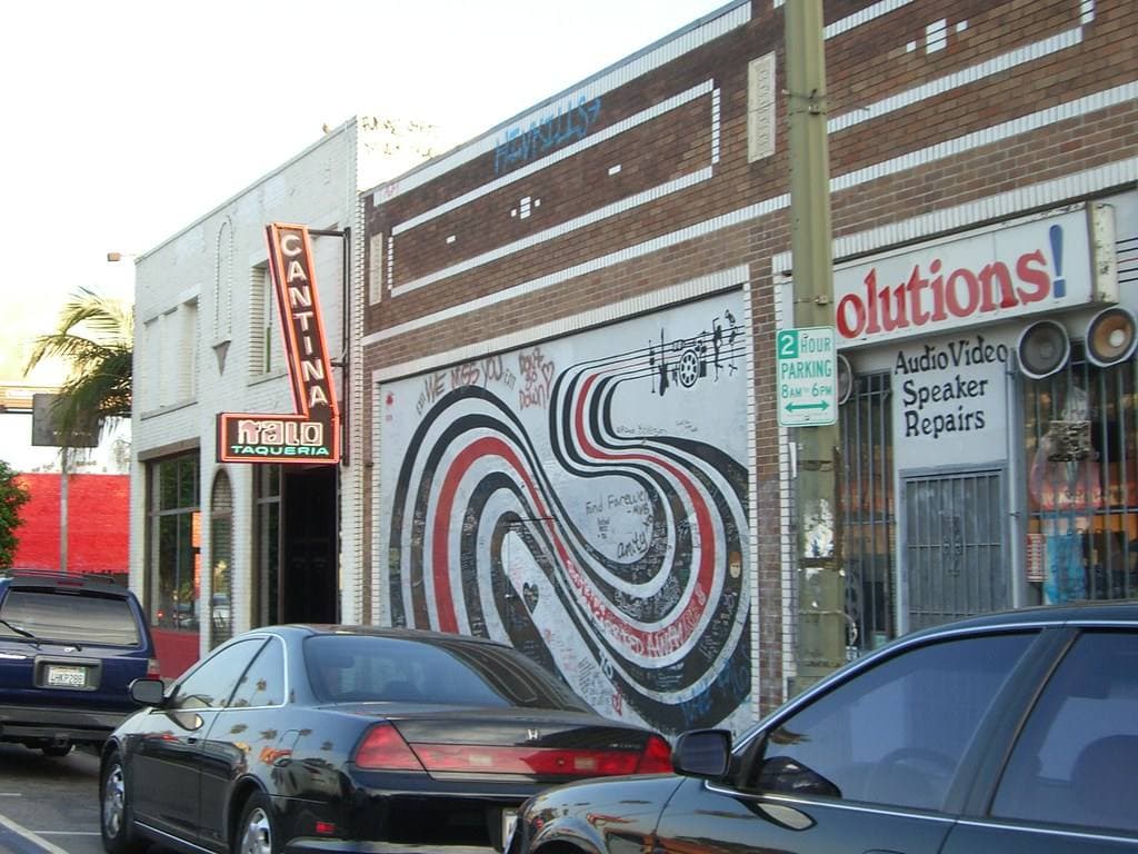 A brick shopfront on Sunset Boulevard with a mural of black, red and white looping bands beside a sign reading Solutions Audio Video Speaker Repairs