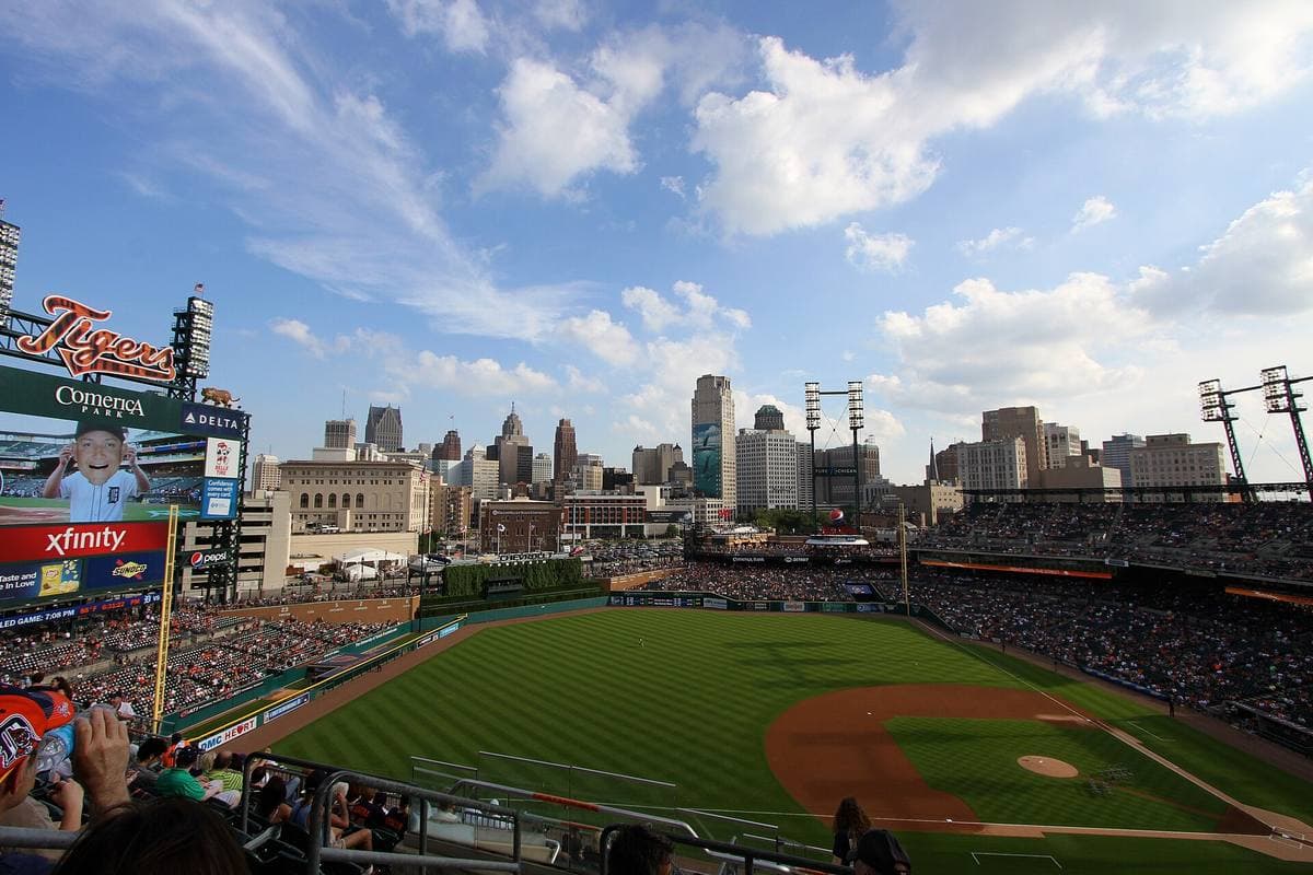 A baseball park full of people in the evening with Detroit towers rising behind
