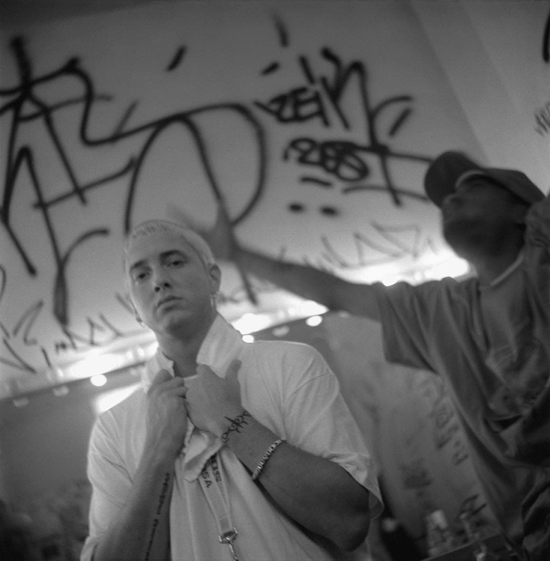 Eminem, bleached hair, white T-shirt, a towel round his neck, looking into the camera backstage under a graffiti-covered wall
